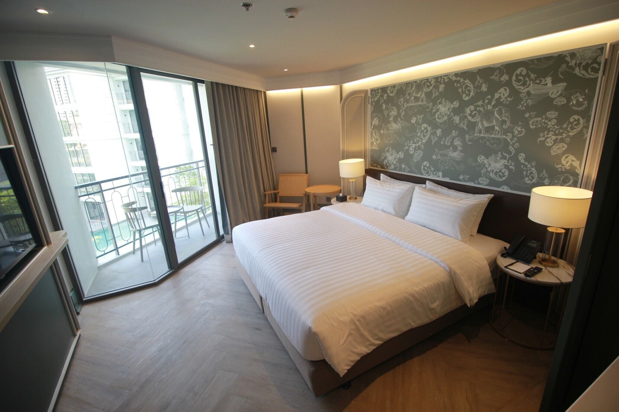 Une chambre d’hôtel moderne avec un grand lit, deux tables de chevet avec des lampes, une petite table ronde et un balcon avec une porte vitrée.