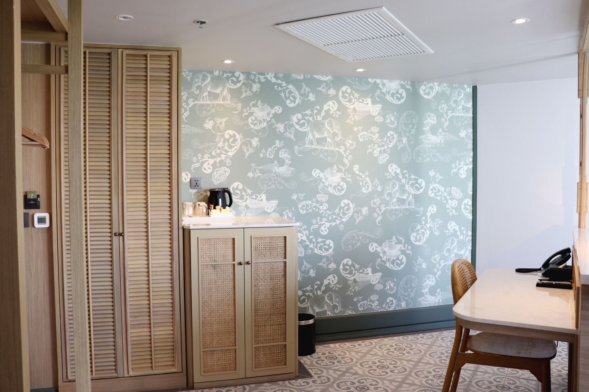 A room with a wooden wardrobe, a small kitchenette with a coffee maker, a patterned wall, and a dining table with chairs.