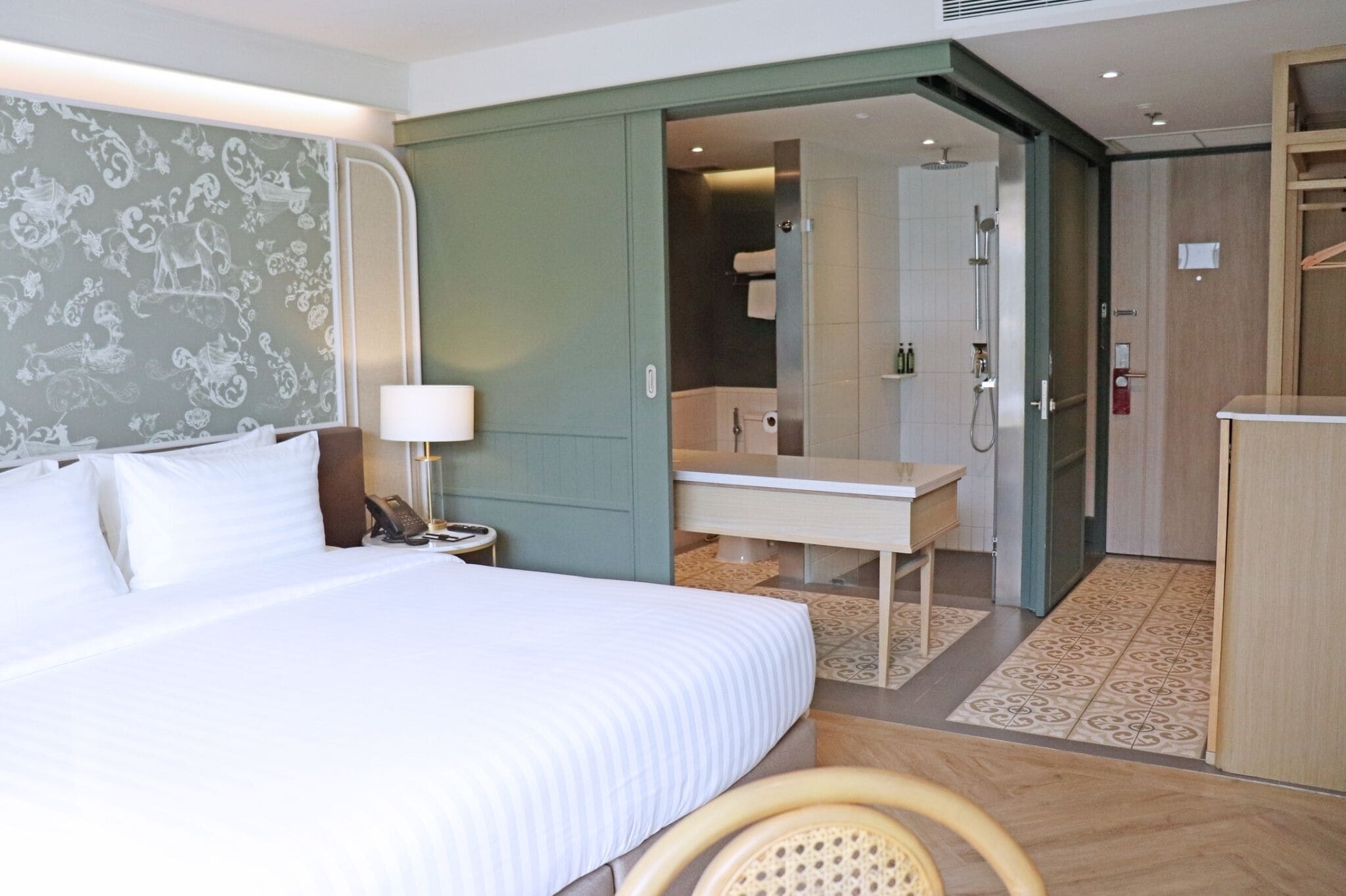 A hotel room with a bed, bedside table, lamp, and a bathroom visible through an open door.