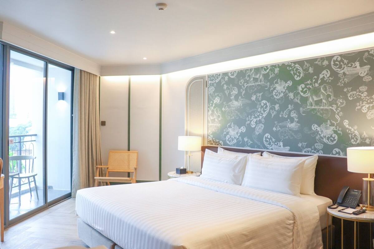 A hotel room with a large bed, two bedside tables with lamps, a chair, and a balcony with a table and chairs.