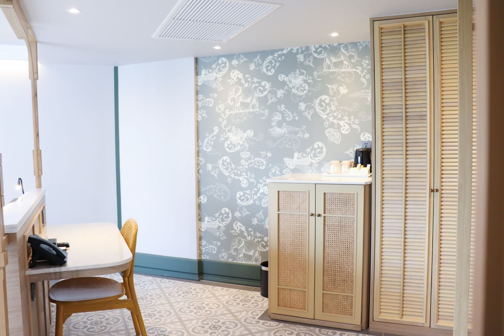 A room with a patterned wallpaper, a wooden cabinet, a small table with a chair, and a wardrobe.