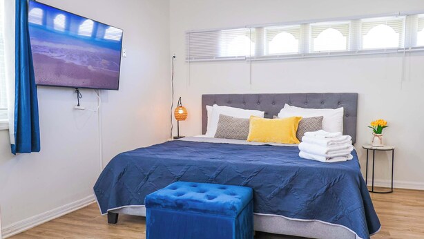 Un dormitorio con una cama, un televisor montado en la pared, una lámpara de noche y una ventana con persianas.