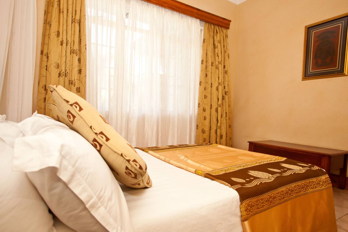 A hotel room with a bed, a wooden nightstand, a framed picture on the wall, and sheer curtains with a floral pattern.