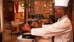 A chef in a white uniform cooking in a kitchen with brick walls and a dining area visible in the background. - Mosa Court Apartments (Kampala)