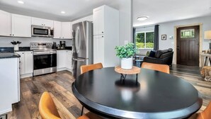 A modern kitchen with stainless steel appliances, a dining area with a round table, and a view of the outdoors through a window. - Bear Walk Cottage in Black Mountain (Black Mountain)