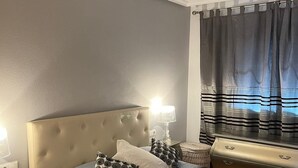 Une chambre à coucher avec une tête de lit capitonnée, un lit avec une literie grise, deux tables de chevet avec des lampes, un ventilateur de plafond et une fenêtre avec des rideaux.