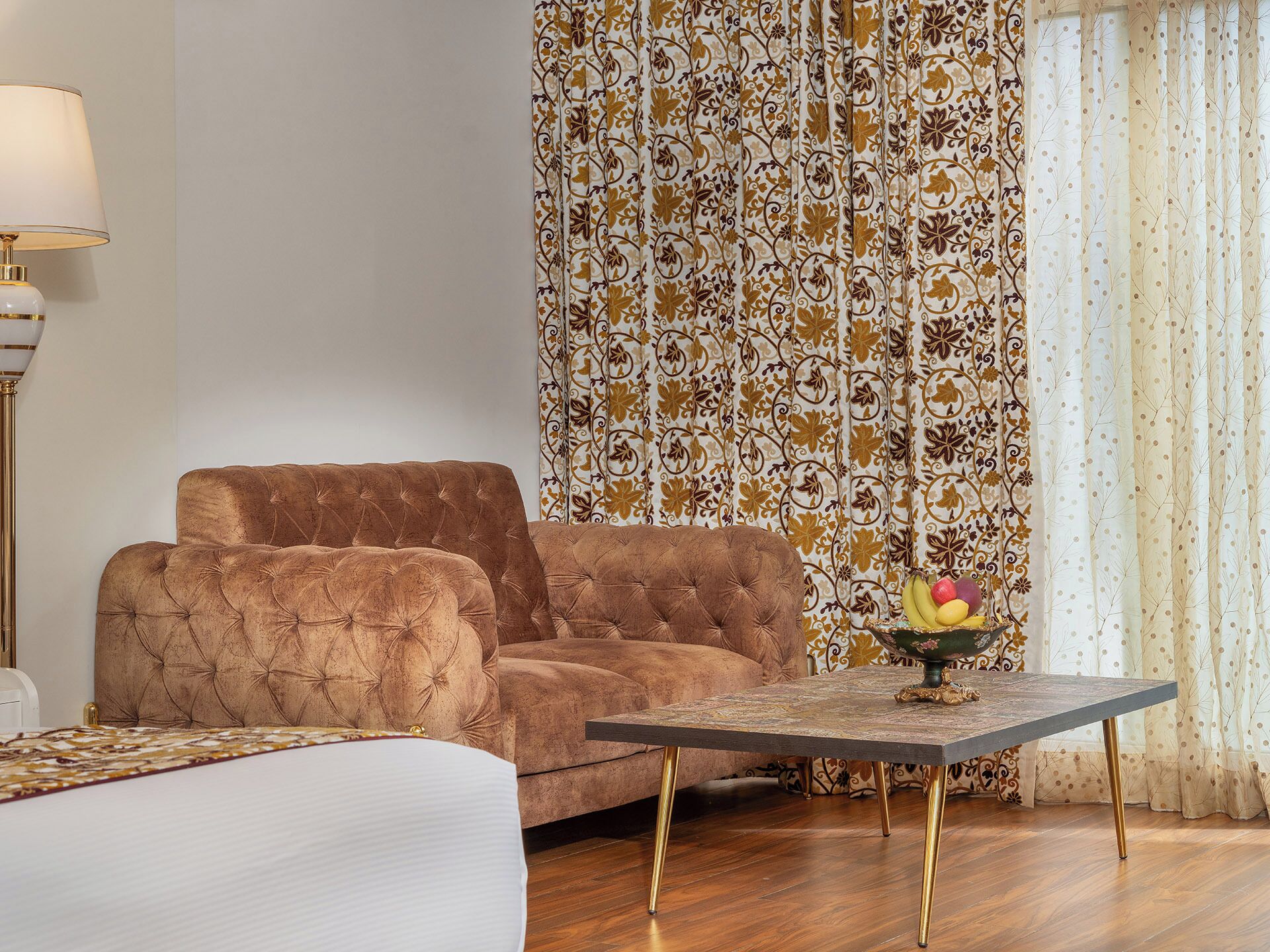 A room with a tufted brown sofa, a small round table with a fruit centerpiece, and patterned curtains.