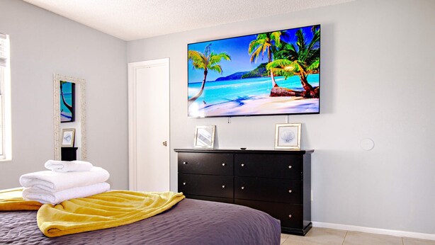 Ein Schlafzimmer mit einem Bett, einem Fernseher an der Wand, einem Schminktisch und einem gerahmten Bild.
