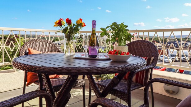A round table with wicker chairs, a bottle of wine, and a bowl of strawberries on a balcony with a view of parked cars and greenery.