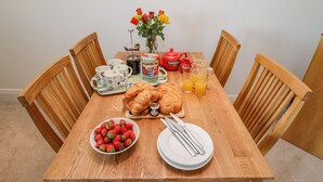 A wooden dining table with a croissant, a bowl of strawberries, a teapot, cups, and a pitcher of orange juice.