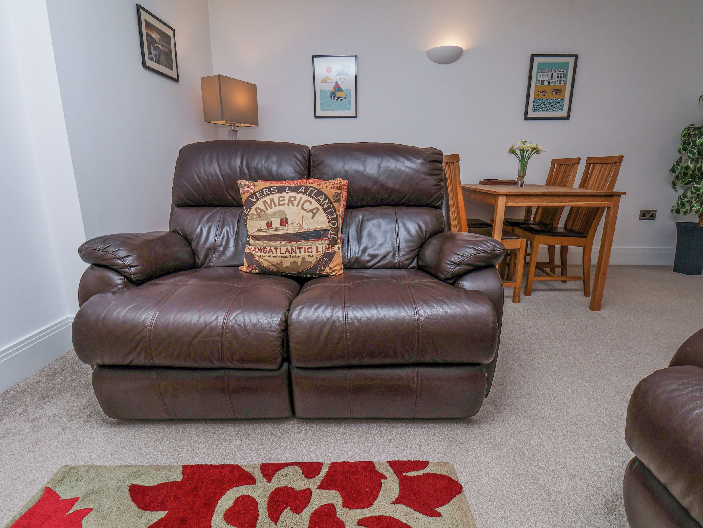 A brown leather sofa with a cushion featuring the Transatlantic Line logo, a wooden dining table with chairs, and a small potted plant.