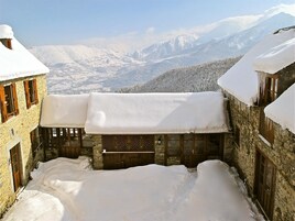 Un patio cubierto de nieve con edificios de piedra, contraventanas de madera y una vista de montañas nevadas al fondo.