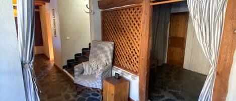 A stone-floored room with a checkered chair and a wooden side table, a wooden lattice wall, and a doorway with curtains.