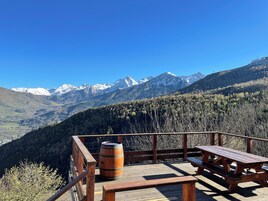 Una terraza de madera con un banco y un barril, con vistas a un paisaje montañoso con picos nevados y un bosque denso.