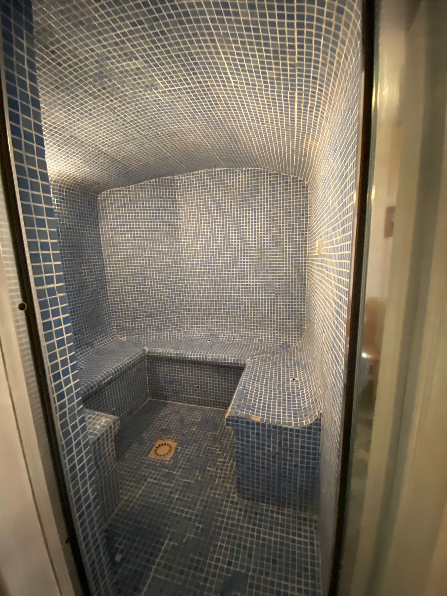 Un área de ducha pequeña y cerrada con paredes y suelo de azulejos azules, que incluye un banco en esquina y un desagüe.
