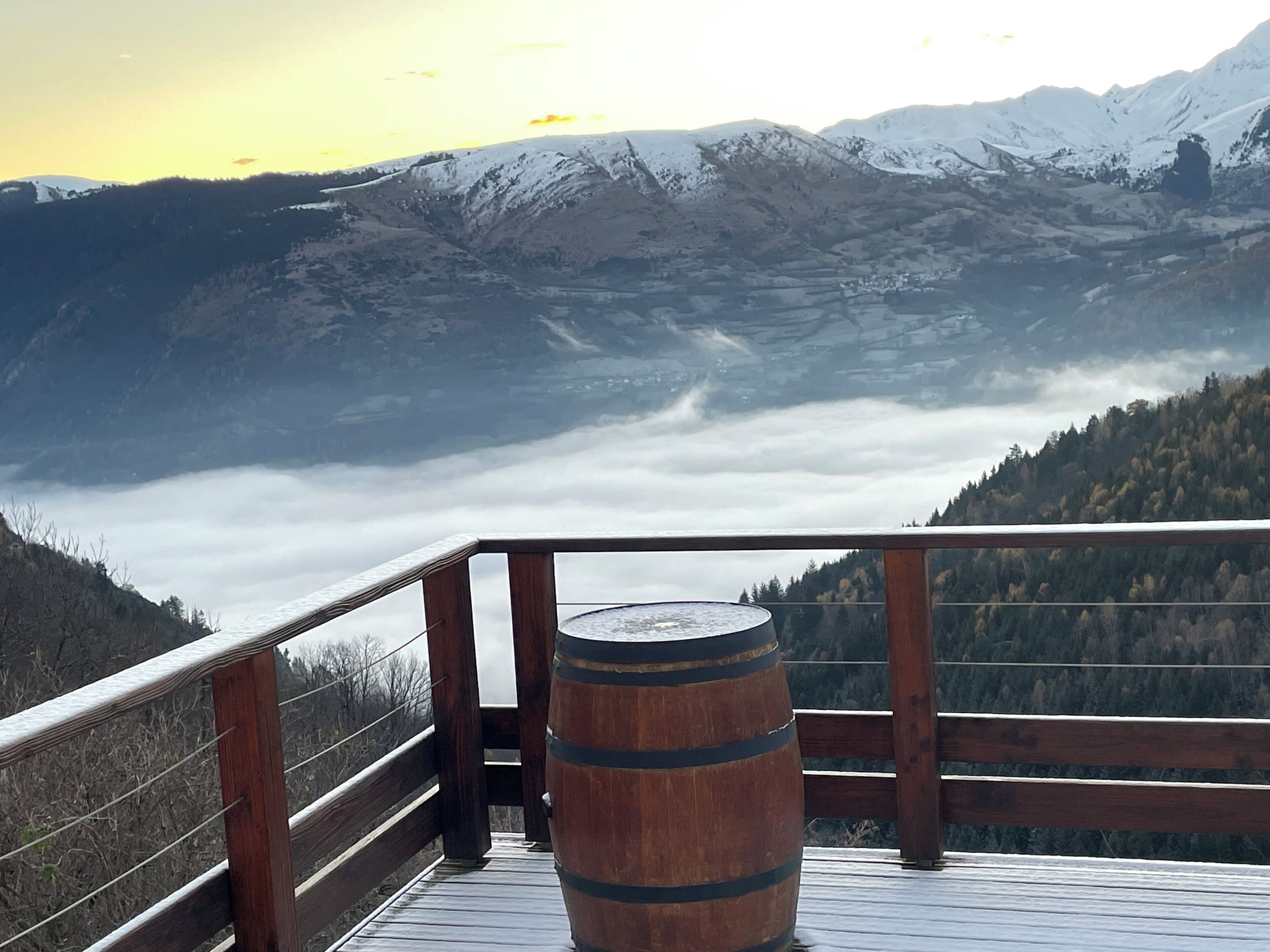 A wooden deck with a barrel, overlooking a valley with snow-capped mountains and a forested area.