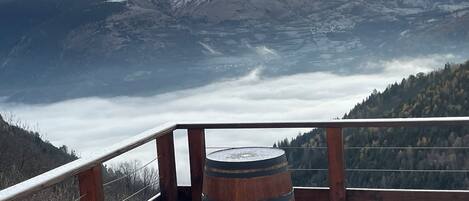 A wooden deck with a barrel, overlooking a valley with snow-capped mountains and a forested area.