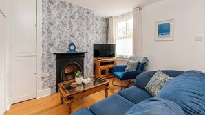 A living room with a blue sofa, a wooden coffee table, a fireplace, a television, and a window with curtains. - Magdala Cottage -Village Location- Dog Friendly (Broadstairs)