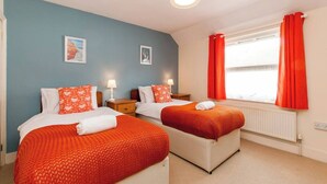 A hotel room with two beds, each with a red blanket and matching pillows. There are bedside tables with lamps, a window with red curtains, and a radiator. - Magdala Cottage -Village Location- Dog Friendly (Broadstairs)