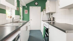 A kitchen with white cabinets, a stainless steel sink, a clock on the wall, and a window with a view of greenery. - Magdala Cottage -Village Location- Dog Friendly (Broadstairs)