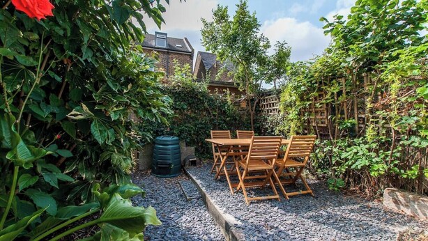 A garden patio with a wooden table and chairs, surrounded by dense foliage and a brick wall. - Magdala Cottage -Village Location- Dog Friendly (Broadstairs)