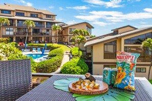 A dining table with a tropical-themed meal, a bag of chips, and cans of soda, overlooking a pool and hotel buildings. - MA 1-310 AC, Fantastic Views, 2 b Loft, 2 1/2 Bath (Koloa)