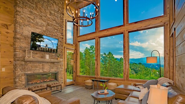 A cozy living room with a stone fireplace, leather sofas, and large windows offering a scenic view. - 🐻Gatlinburg Mountain Romance: Secluded, Honeymoon, Outdoor Fireplace, Hot Tub🏞 (Gatlinburg)