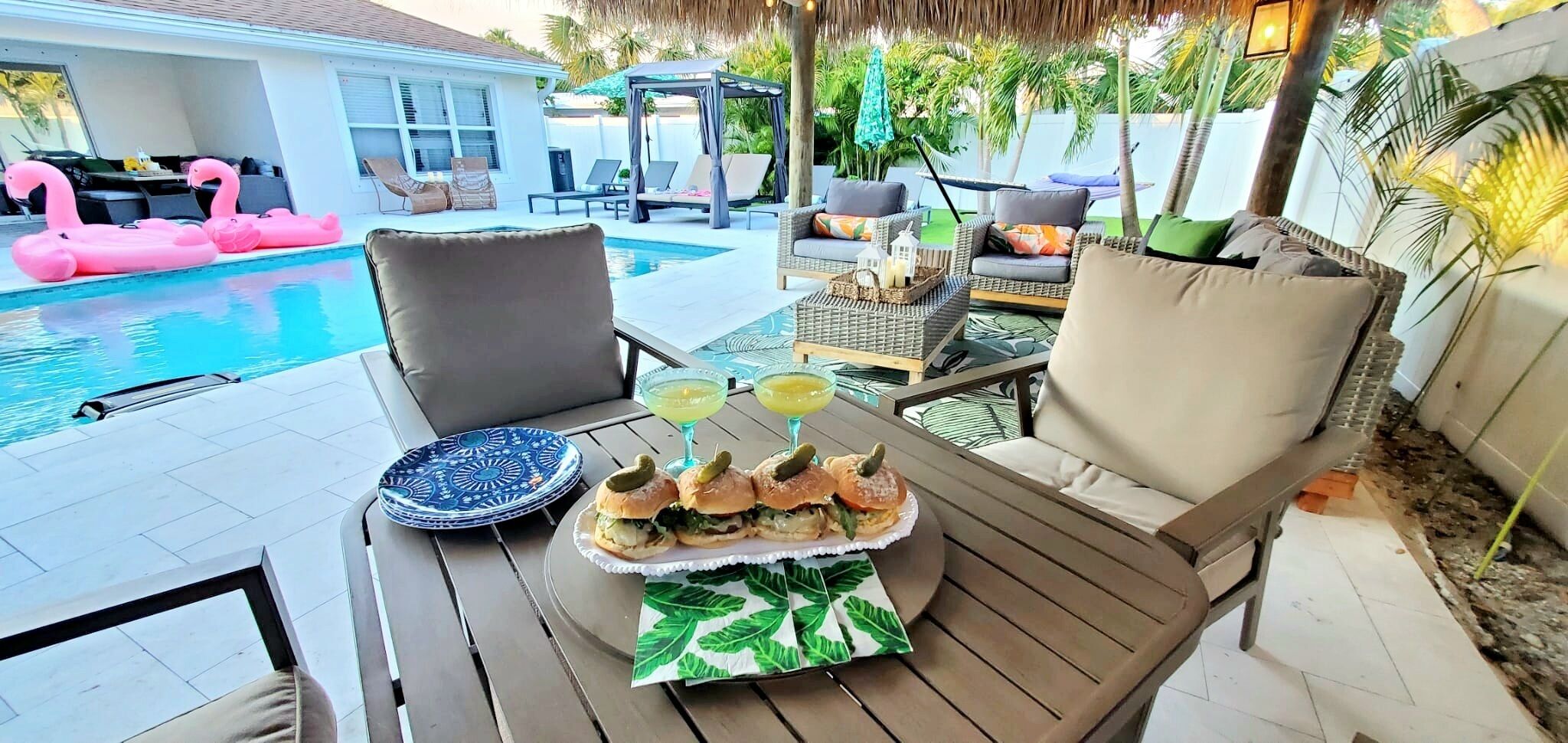 Spiseområde ved poolen med et bord dækket til et måltid, inklusive sandwiches og drikkevarer, samt en pool med lyserøde flamingoer.