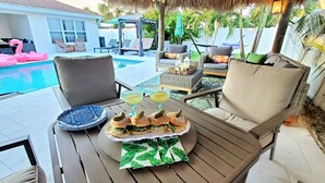 A poolside dining area with a table set for a meal, including sandwiches and drinks, and a pool with pink flamingos.