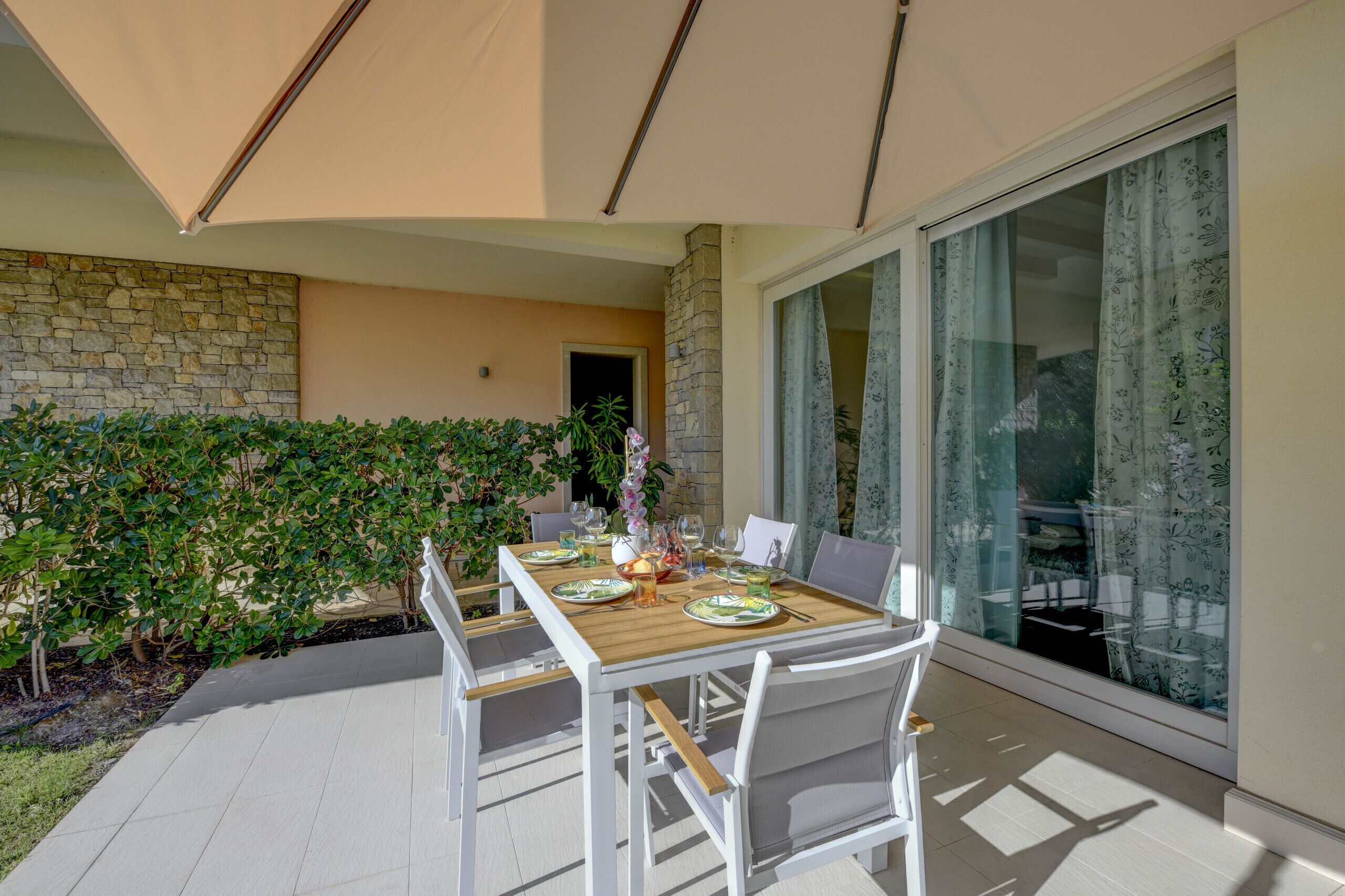 An outdoor dining area with a wooden table and white chairs, surrounded by a stone wall and greenery.
