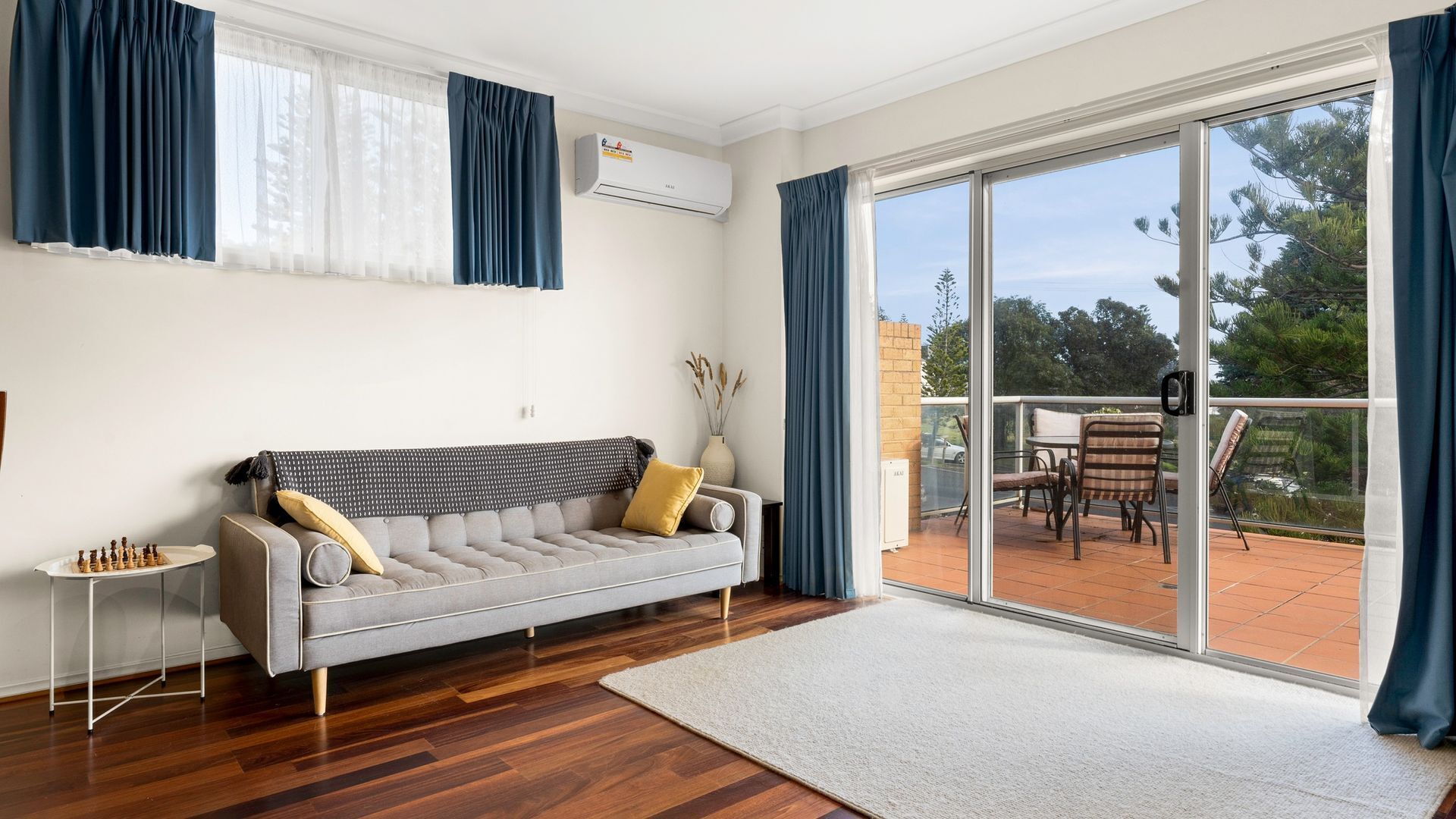 A modern living room with a grey sofa, a chessboard, and a large sliding glass door leading to an outdoor patio.