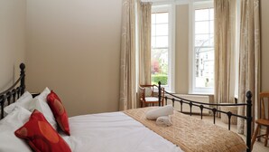 A neatly made bed with a white comforter and red pillows, a wooden headboard, and a view of the outdoors through large windows with curtains. - Magpies (St Leonards-On-Sea)