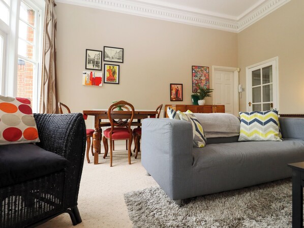 A living room with a grey sofa, a black wicker chair, a dining table with chairs, and a grey rug.