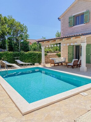 A rectangular swimming pool with a stone deck, surrounded by lounge chairs and a pergola with hanging lights.