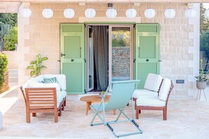 A patio with a wooden table, two wooden chairs, and a metal folding chair. There are green shutters on the doors, a potted plant, and a stone wall.