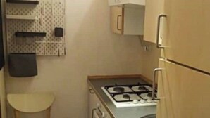 Ett litet, praktiskt kök med en gasspis, en diskho och skåp. Det finns en stol och en väggpanel med krokar på väggen.