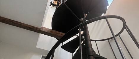 Una escalera en espiral con barandilla de metal, viga de madera y lámpara colgante.
