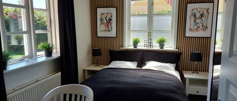 Une chambre à coucher comprenant un lit, deux tables de chevet, une chaise, une fenêtre avec des rideaux et deux plantes en pot.