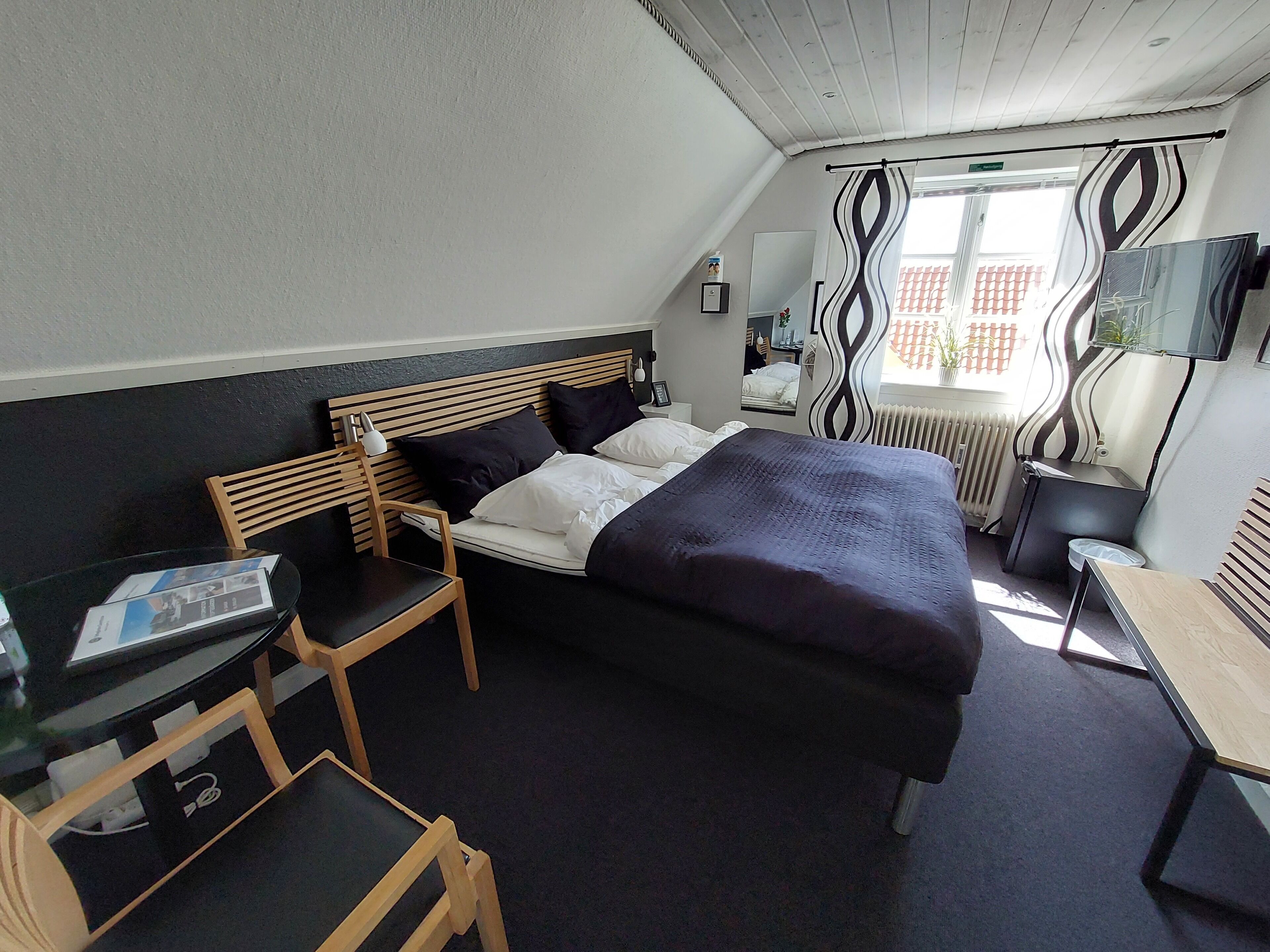 Habitación de hotel con cama, escritorio, silla, televisor y ventana con cortinas.