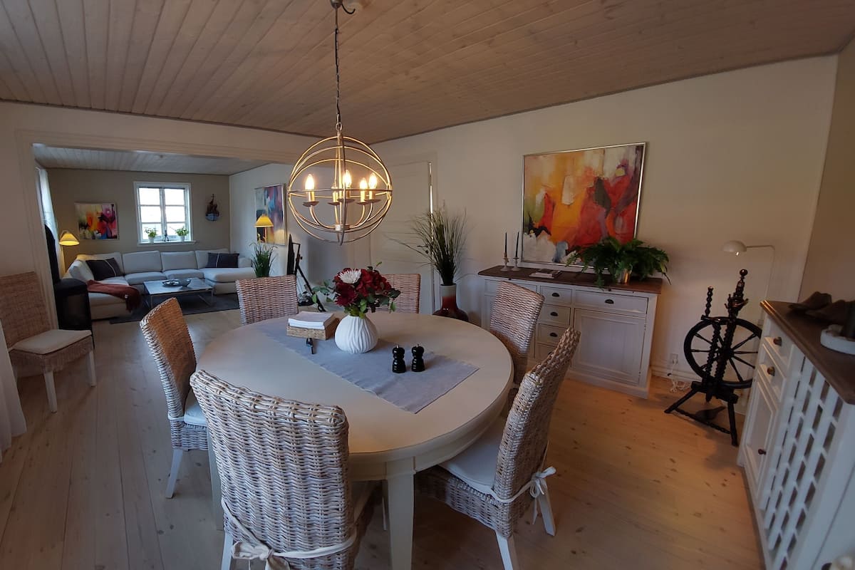 A dining area with a round table, wicker chairs, a chandelier, and a painting on the wall.