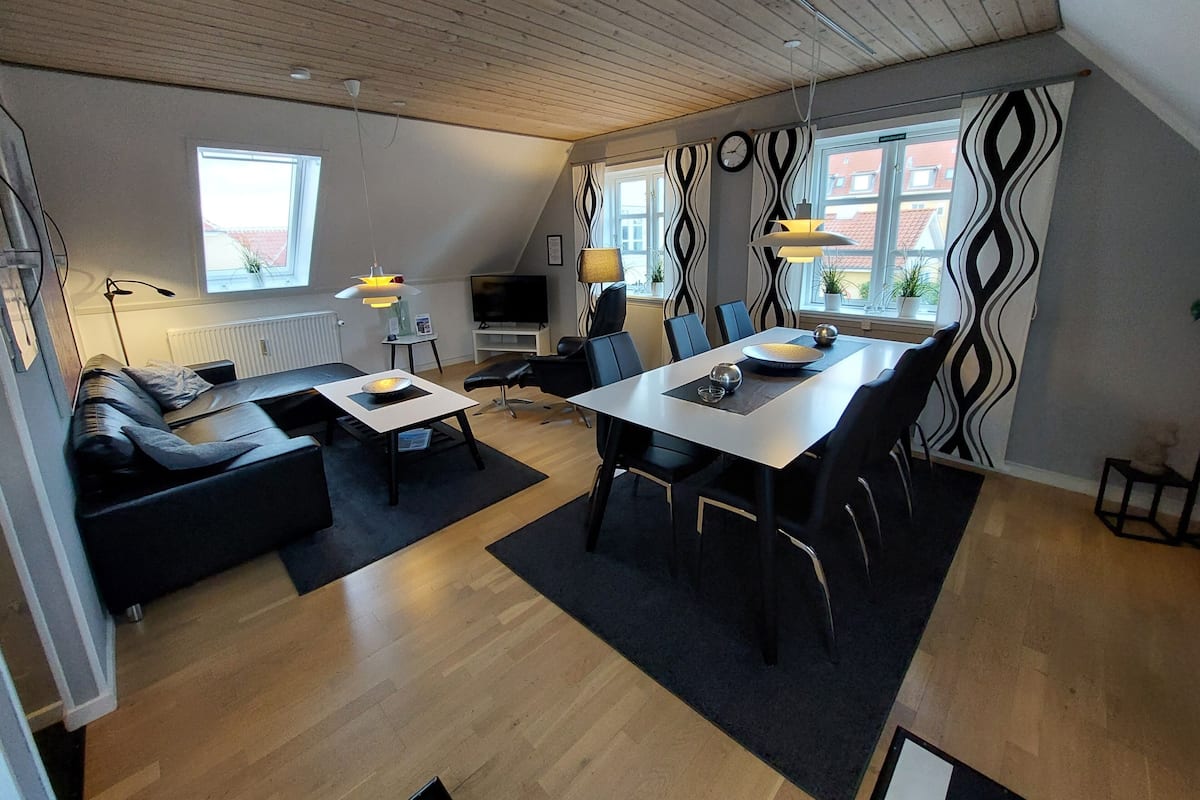A modern living room with a sofa, dining table, and chairs, a TV, and a large window with a view of buildings.