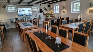 Un restaurante con varias mesas y sillas de madera, un televisor mostrando un menú y una zona de cocina visible al fondo.