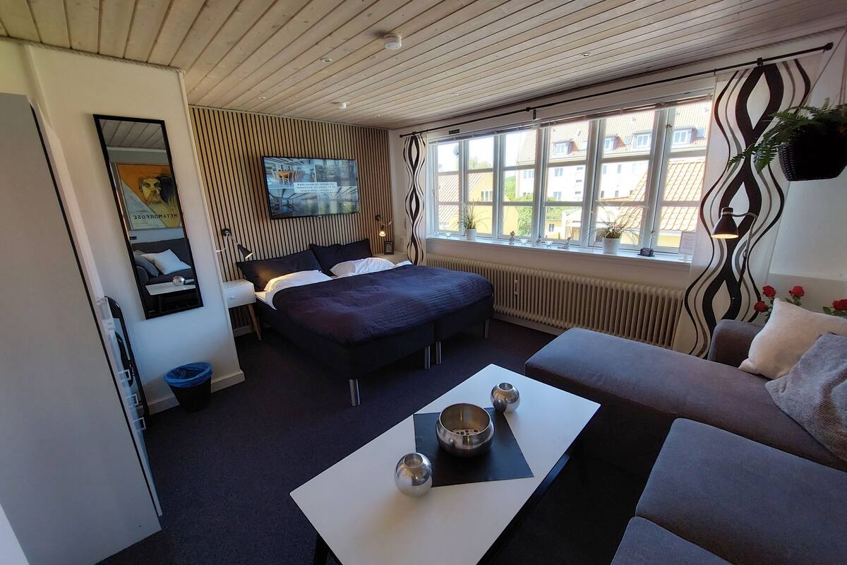 A modern hotel room with a large bed, a sofa, a TV, and a window with a view of buildings.