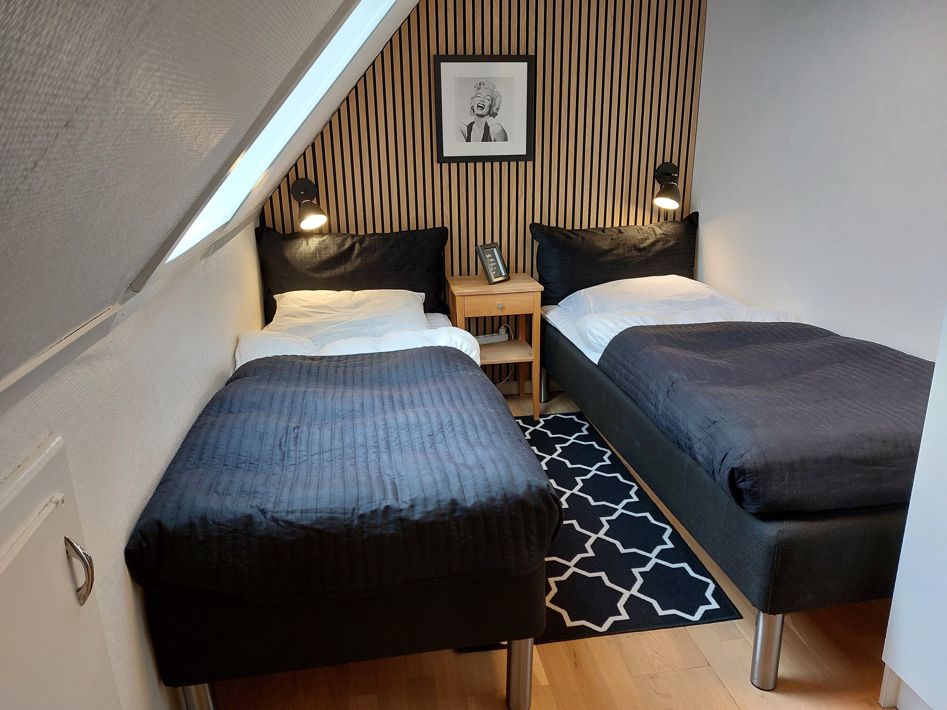 Une chambre à coucher de taille modeste, avec deux lits, un mur recouvert de lambris en bois, un tableau encadré et une petite table de chevet avec une lampe.