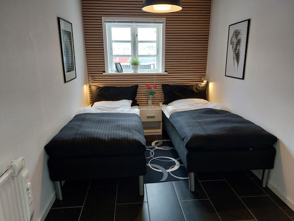 A small, modern bedroom with two single beds, a window with blinds, a small table with a plant, and a radiator.