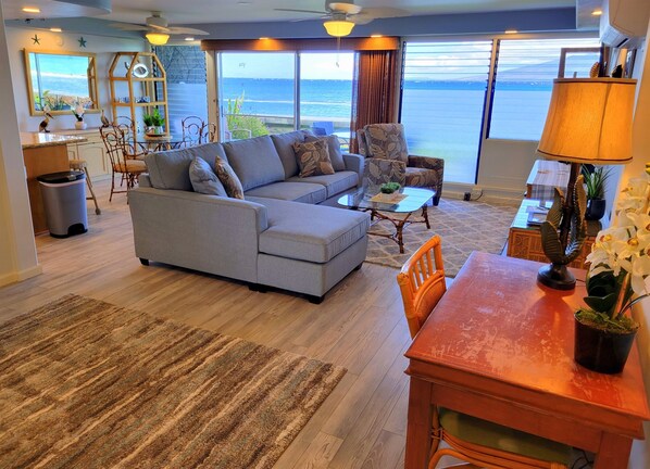 A living room with a large gray sectional sofa, a wooden dining table with chairs, a coffee table, and a view of the ocean through large windows. - Exquisite Direct Sunset & Oceanview 4 Miles! Pool (Lahaina)
