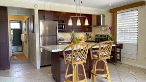 A modern kitchen with dark wooden cabinets, stainless steel appliances, a central island with bar stools, and a window with blinds. - Serene Private Ocean View  AC  Pool (Lahaina)