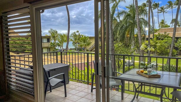 A balcony with a table and chairs, overlooking a garden with palm trees and a view of the ocean. - Serene Private Ocean View  AC  Pool (Lahaina)