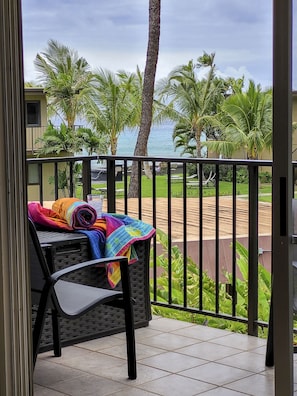 A balcony with a chair and a colorful blanket, overlooking a beach with palm trees and a view of the ocean.