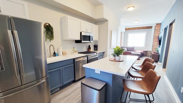 A modern kitchen with stainless steel appliances, white cabinetry, and a central island with bar stools. - Craft District Pet-Friendly Condo by Heirloom (Duluth)